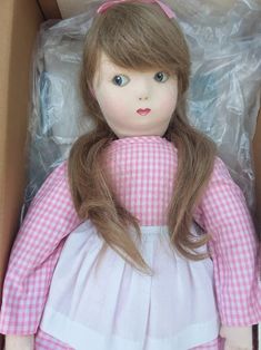 a doll in a box with its hair pulled back and wearing a pink checkered dress