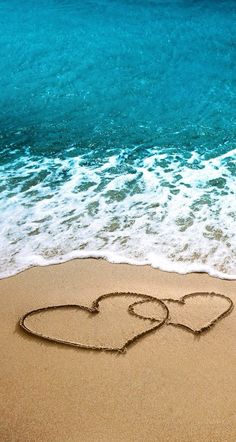 two hearts drawn in the sand on a beach with blue water and waves behind them