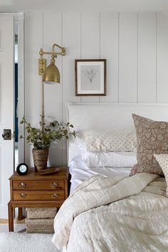 a bed with white sheets and pillows in a bedroom next to a wooden dresser topped with potted plants