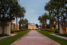 a brick walkway leading to a white house surrounded by palm trees and greenery at dusk