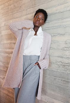 a woman leaning against a wall with her hands on her head, wearing a white shirt and gray pants