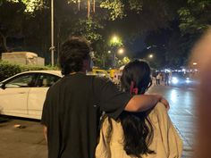 a man and woman walking down the street at night with cars parked in the background