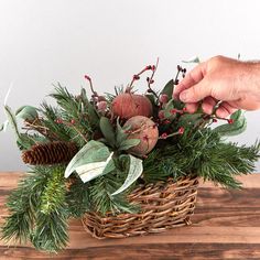 a basket filled with greenery and pine cones on top of a wooden table next to a person's hand