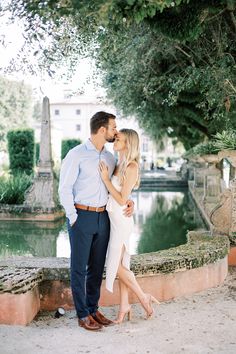 a man and woman standing next to each other in front of a pond with trees