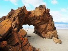 a large rock formation on the beach with a person standing in it's doorway