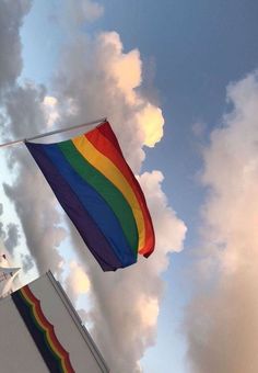 a rainbow flag flying in the sky with clouds behind it and a clock tower below