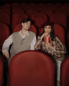 a man and woman sitting in the middle of an empty auditorium with red seats, one holding a drink