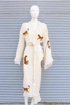 a white robe with cats on it and fringes around the collar, is displayed in front of a metal wall