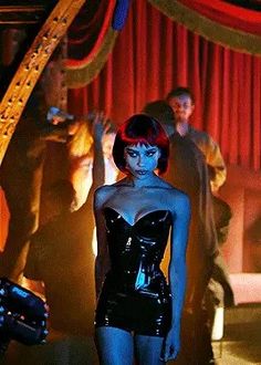 a woman with blue skin and black latex is standing in front of red curtains