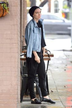 a woman in black pants and a denim jacket is standing on the sidewalk near a bench