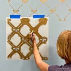 a woman is painting a wall with gold paint and stencils in front of a blue background