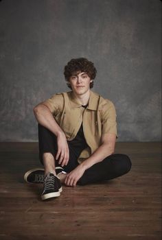 a young man sitting on the floor with his legs crossed, wearing black pants and a tan shirt