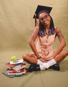 a woman sitting on the ground with books and an apple in front of her, wearing a graduation cap