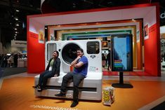 two men sitting on top of a washing machine in front of a tv screen at an exhibition