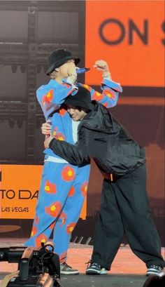 two men in colorful clothing are hugging each other on stage with an orange and black sign behind them