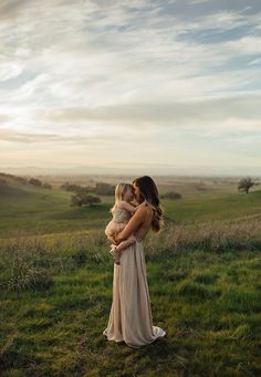 a woman holding a baby in her arms while standing on top of a lush green field