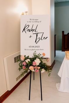 a welcome sign with flowers and greenery in front of a white table clothed backdrop