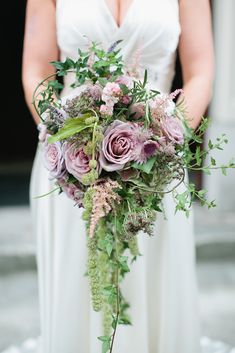 a woman in a white dress holding a bouquet of purple flowers and greenery on her wedding day