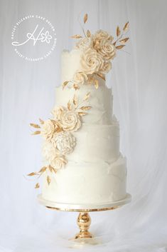 a three tiered cake with flowers on top and gold leaves around the edges, against a white backdrop