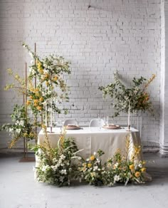 an arrangement of flowers and greenery on a table in front of a brick wall