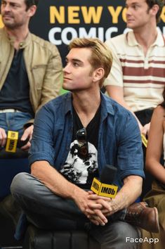 a man sitting in front of a microphone with other people behind him at a comic convention