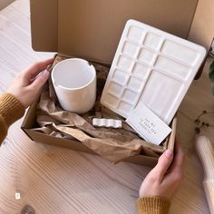 two hands are holding a box with a white cup and saucer in it on a wooden table