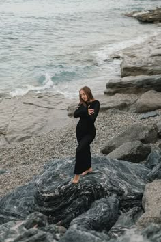 a pregnant woman standing on rocks near the ocean with her arms crossed and looking at the camera