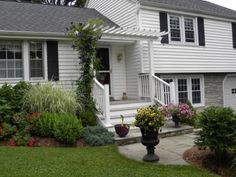 a white house with black shutters and flowers in the front yard on a cloudy day