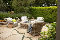 a stone patio with chairs and fire pit surrounded by greenery in the back yard