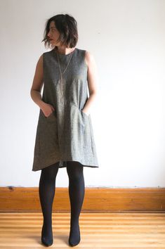 a woman standing in front of a white wall wearing black tights and a gray dress