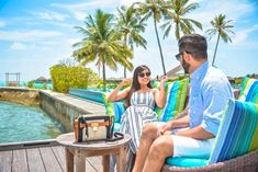 a man and woman sitting on top of a wooden deck next to the ocean in front of palm trees