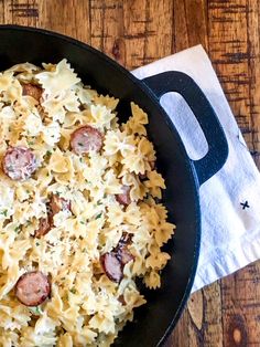 a skillet filled with pasta and sausages on top of a wooden table next to a napkin