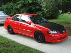 a red car parked on the side of a road next to a silver car in front of some trees