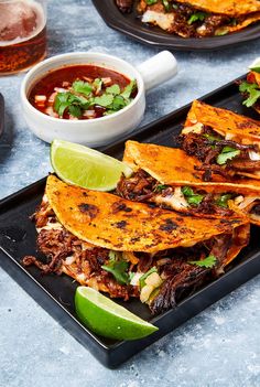 some tacos are on a black plate with lime wedges and salsa in the background