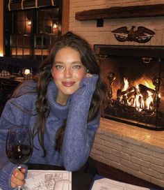 a woman sitting at a table with a glass of wine in front of a fire place