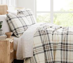 a bed with white and black plaid comforter next to a wooden nightstand in front of a window