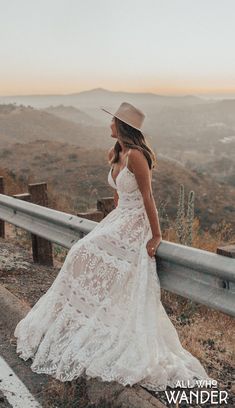 a woman in a white dress and hat leaning on a fence looking at the mountains