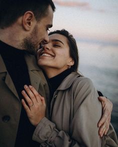 a man and woman standing next to each other near the ocean with their arms around each other