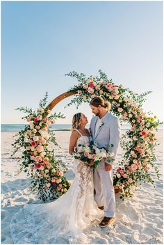 a bride and groom standing in front of a floral arch on the beach at sunset