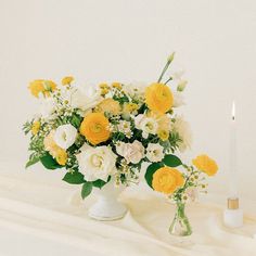 a white vase filled with yellow and white flowers next to a lit candle on a table