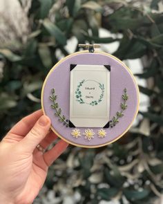 a hand holding up a small embroidery hoop with flowers and leaves on it, in front of a plant