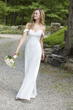 a woman in a white wedding dress walking down a gravel road holding a flower bouquet