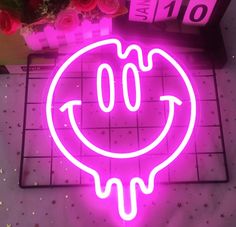 a pink neon sign with a smiley face on it's side next to flowers