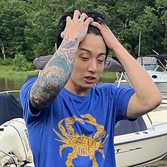 a young man with tattoos on his arms standing next to a boat in the water