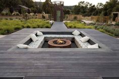 an outdoor fire pit surrounded by wooden decking and seating with white pillows on it