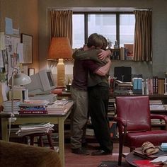 two people hugging each other in a living room with lots of books on the table