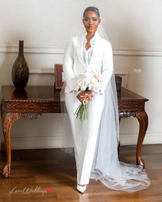 a woman in a white suit and veil standing next to a table with flowers on it