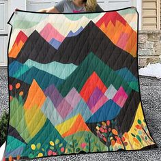 a woman holding up a colorful quilt on the side of a house with mountains in the background