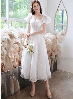 a woman standing in front of a window wearing a white dress and holding a bouquet