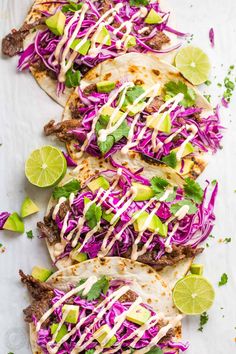 three tacos with shredded cabbage, limes and cilantro on a sheet of paper